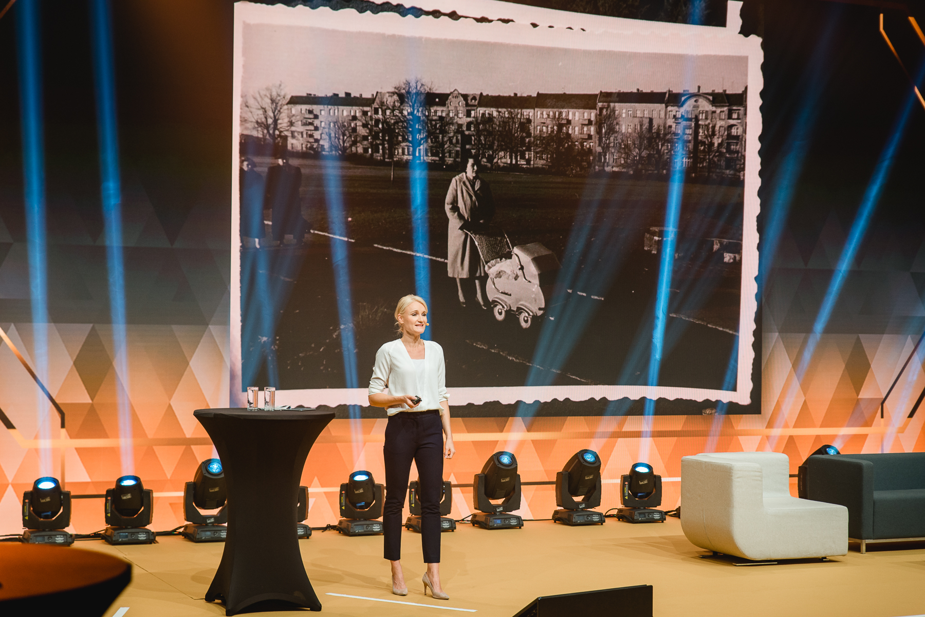 Henriette Hochstein-Frädrich on a large corporate stage, speaking in front of a historic black-and-white photo backdrop under dramatic lighting