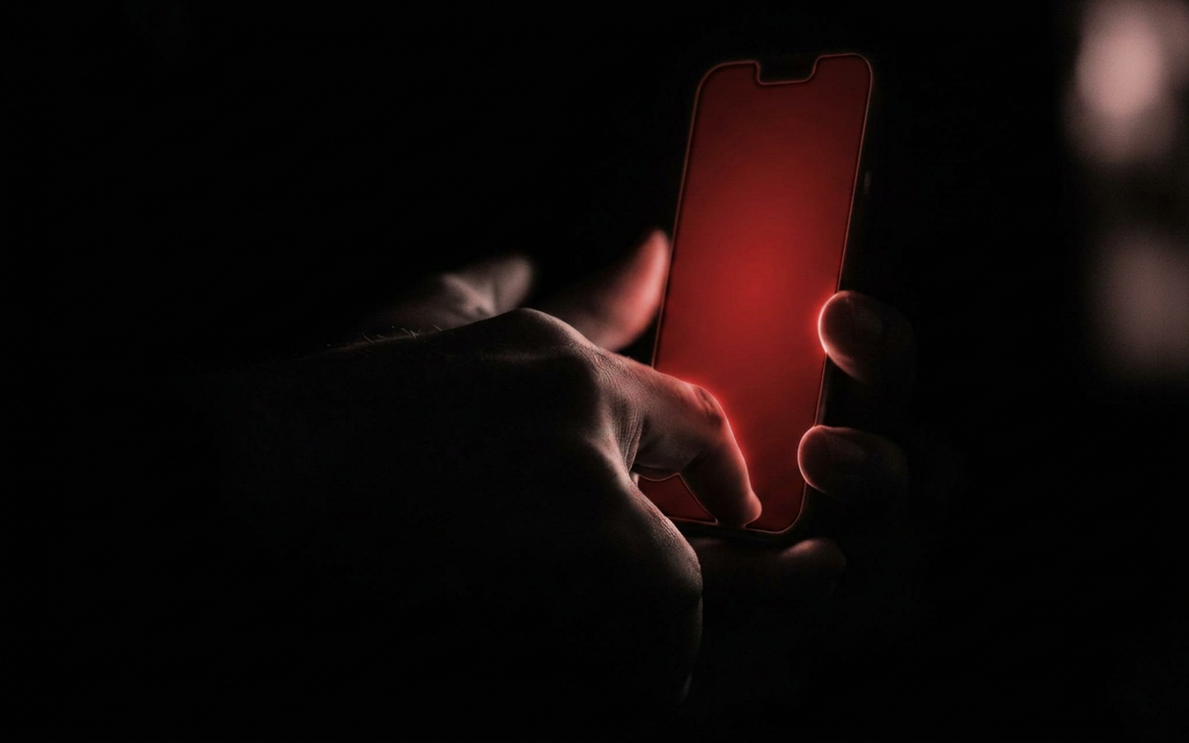 Editorial black and white photograph of an adult hand reaching toward a glowing smartphone in the dark, the screen light catching the fingertips