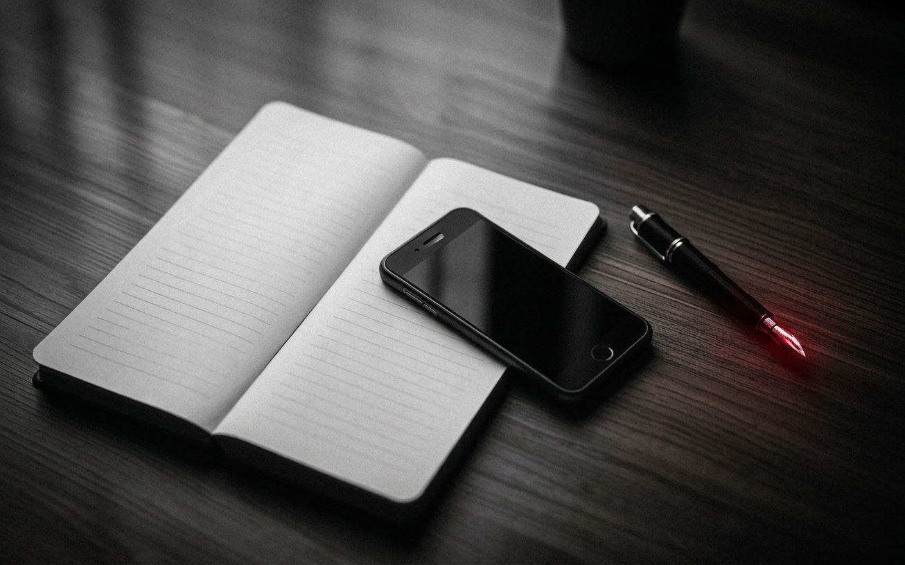 Editorial black and white photograph of a smartphone lying face-down on an empty wooden desk next to an open analog notebook and a fountain pen, soft morning light